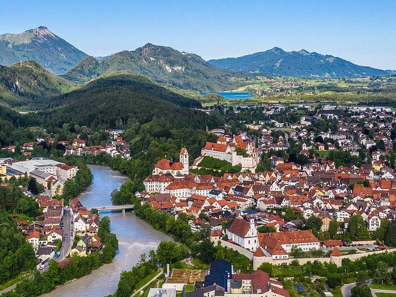 Drohnenaufnahme der Stadt Füssen. Der Lech durchzieht die Stadt. Im Hintergrund ist ein See und Berge zu sehen.